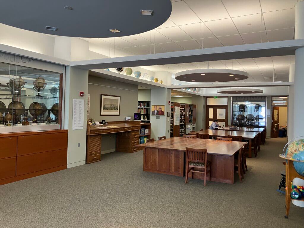 Image of the interior of the Osher Map Library Reading Room. Three large tables are central while library book stacks, globe displays, and a computer are visible along the perimeter of the room.